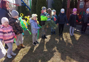 Dzieci stoją w ogrodzie przedszkolnym, trzymają marzannę i gaik
